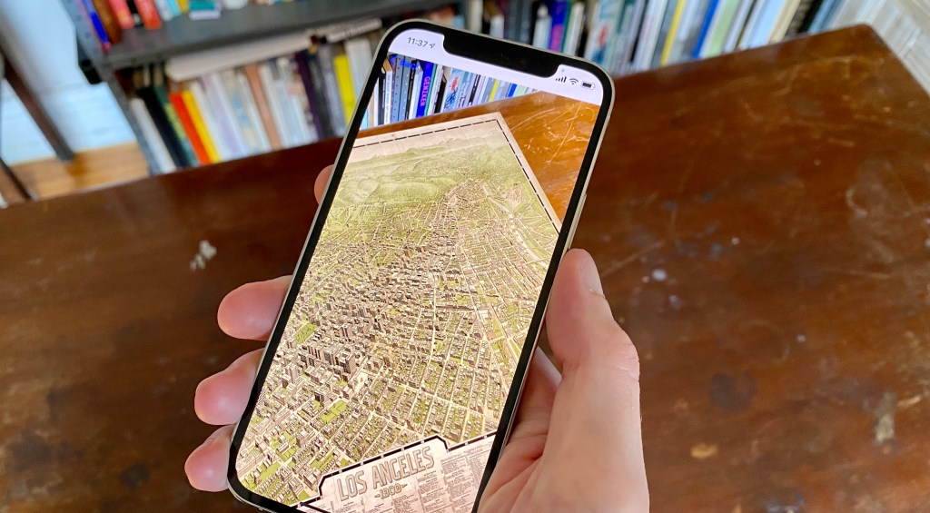 A hand holds an iPhone above a desk. Nothing is on the desk surface. The phone screen displays an archival map superimposed on the desk surface.